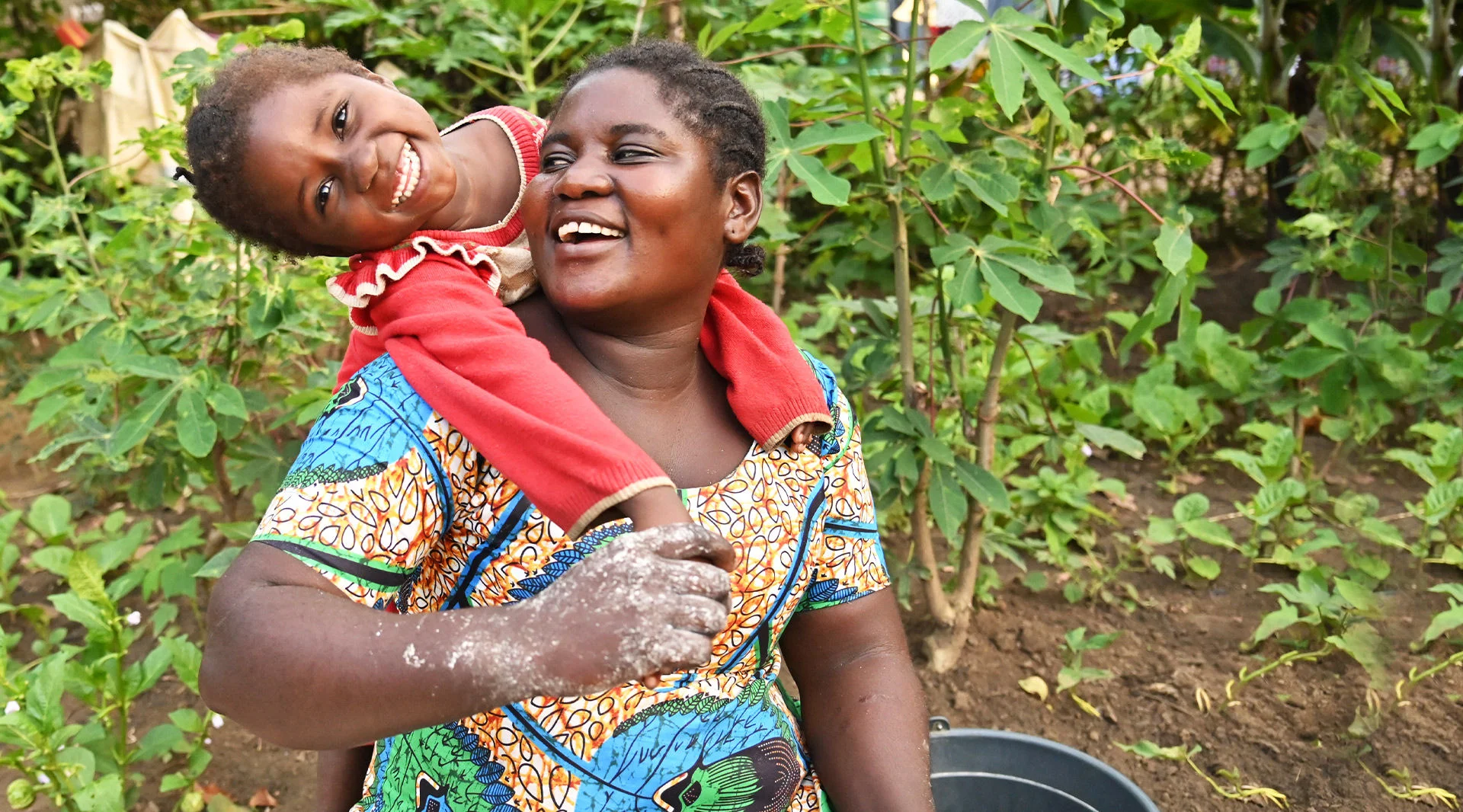 A daughter drapes her arms over her mother’s shoulders as they smile and laugh. (DRC, 2024)