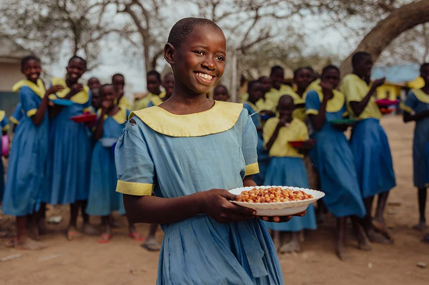 A child holds a plate of food during a World Vision school feeding program, with other children gathered nearby.