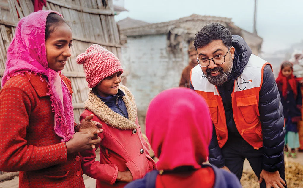 A man wearing a World Vision vest looks down and talks to three children outside their home in Nepal.