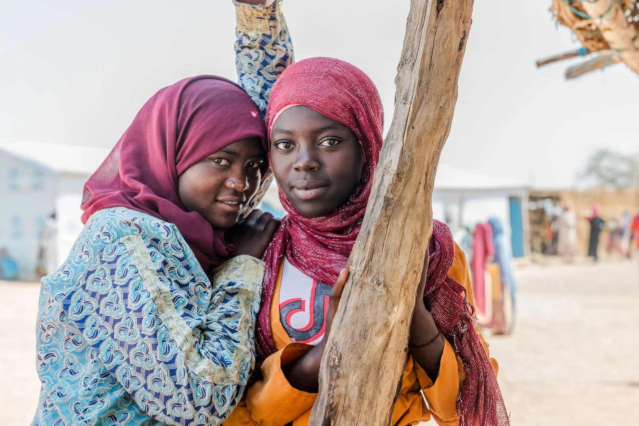 Two young people stand close together beside a wooden post in an outdoor settlement.