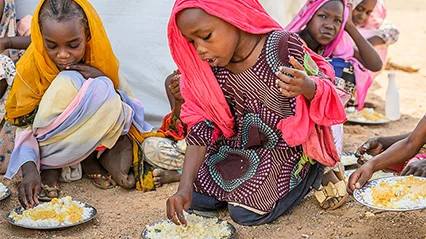 A group of girls wearing headscarves sitting on the ground in a Sudanese refugee camp and eating from plates of donated food. 