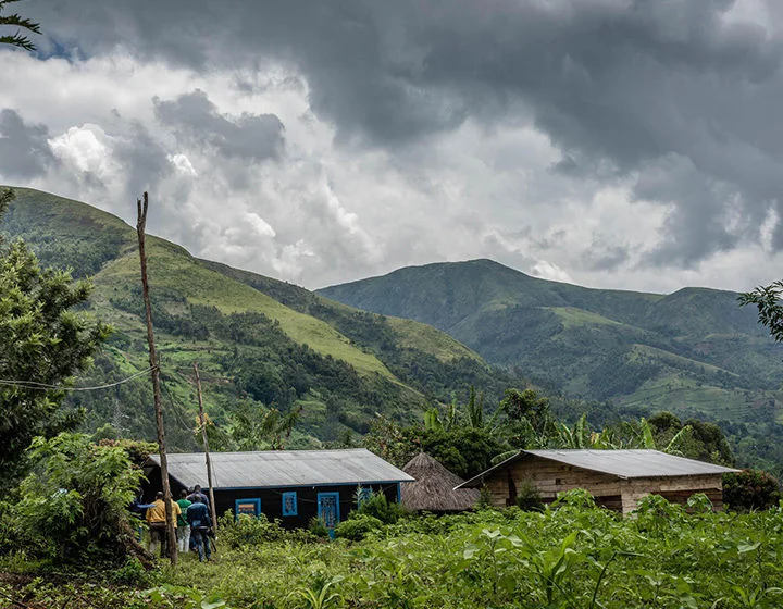 A wide shot image of a green forested area in the Democratic Republic of Congo. In the distance there are people walking towards two large cabins. (Democratic Republic of Congo, 2022)