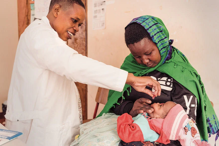 A woman wearing a green headscarf holds a baby as a healthcare worker examines her. (Tanzania, 2025)