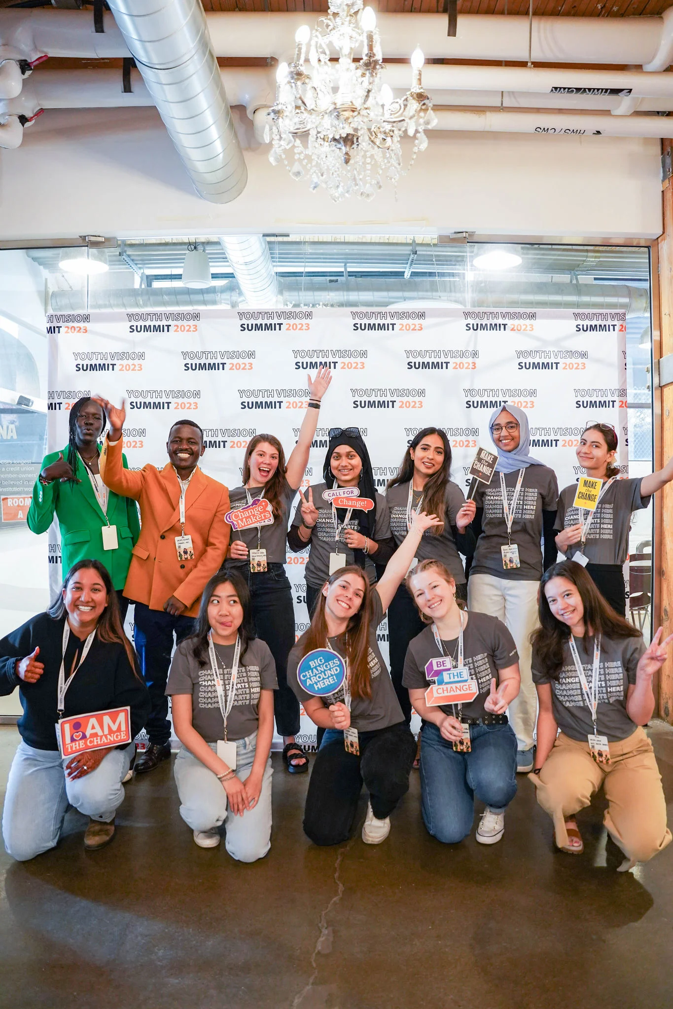 Group of people posing in front of a 'SUMMIT 2023' backdrop, holding signs and wearing lanyards with badges. A chandelier hangs above them.