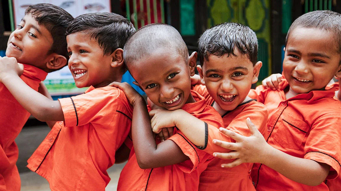 Three young boys wearing orange uniform shirts are hugging each other and smiling.