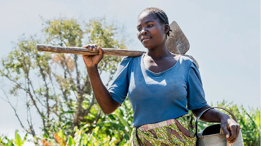 A woman wearing a blue shirt stands in her garden, holding a metal watering can on one arm and a gardening tool on her shoulder.