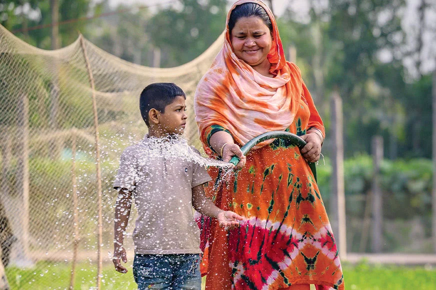 A woman wearing an orange dress waters her vegetables with a young boy beside her. (Bangladesh, 2025)