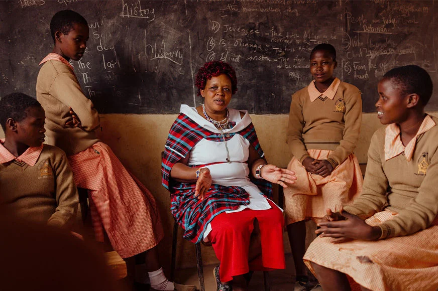 Peninah, an advocate for girls in Osiligi AP Kenya, is sitting in a classroom with her students.