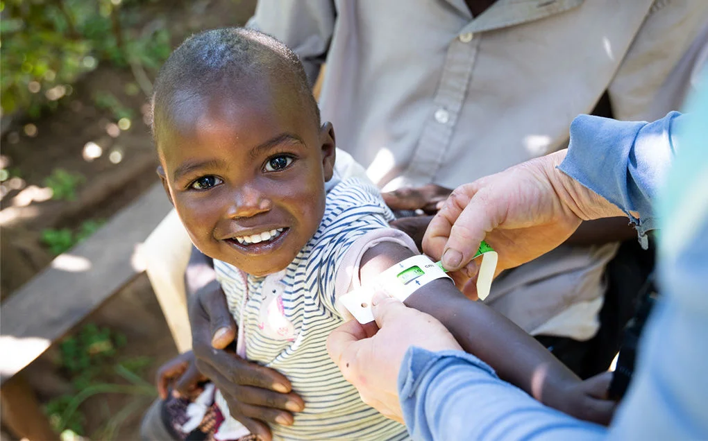 A child smiles while having their arm circumference measured.