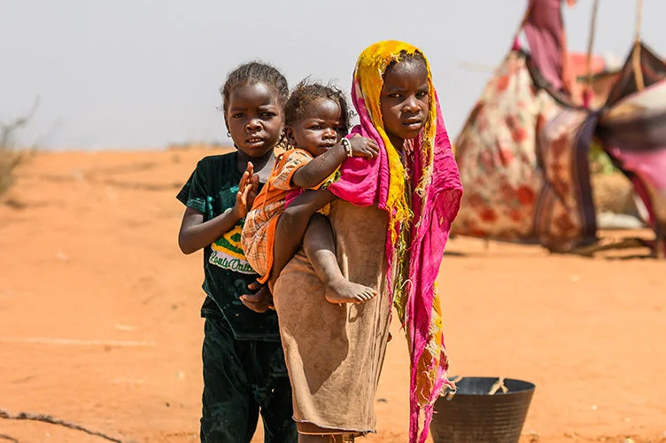 Three Sudanese children stand together on sandy ground in a spontaneous settlement at Adre, Chad.