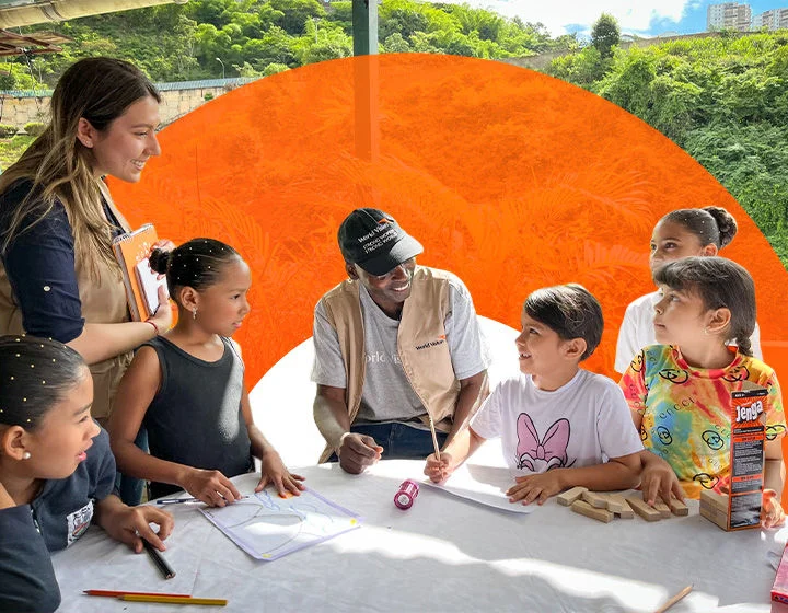 World Vision staff leading outdoor educational activity with children, featuring drawing materials and games on a table in a lush, green setting. 