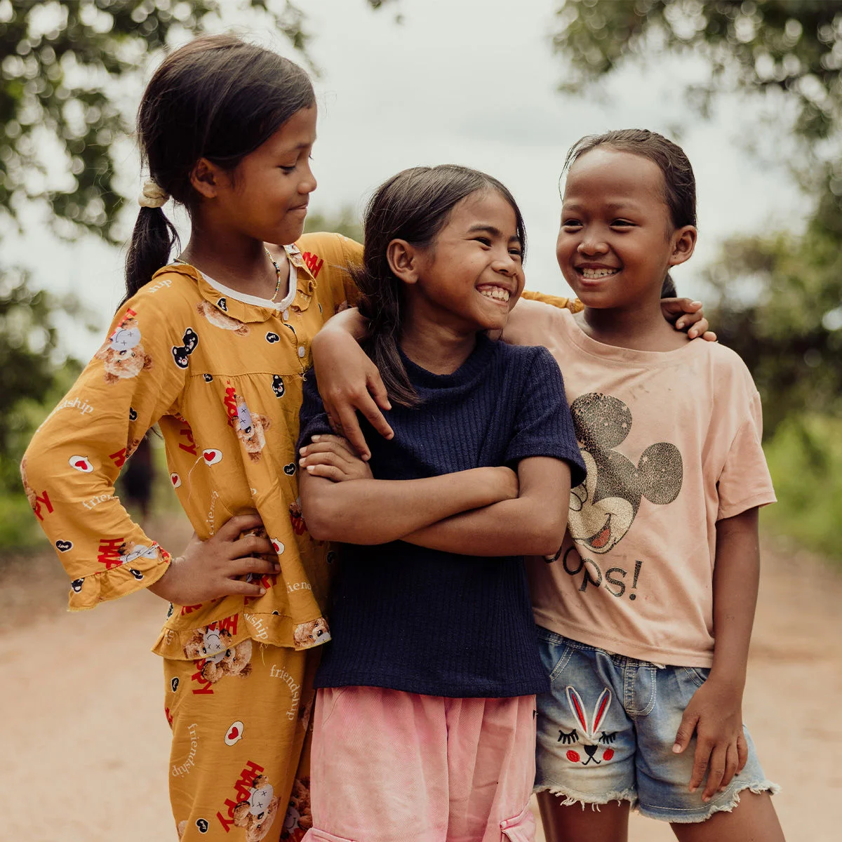 Three young girls standing together on a dirt road with their arms around each other, smiling and surrounded by greenery.
