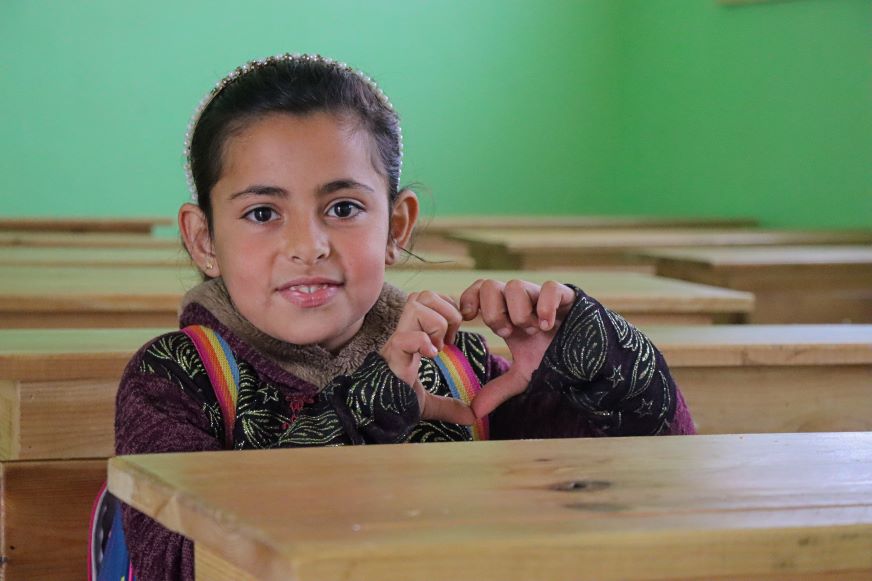 Firyal’s smile and she sits at a school desk and makes a heart symbol with her hands. Syria