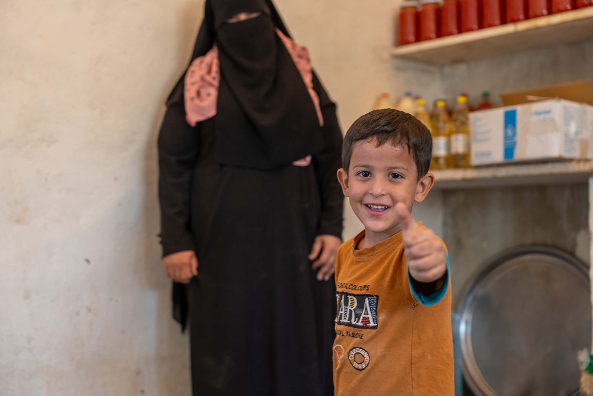 A young boy wearing an orange shirt gives a thumbs up, while his mom stands in the background.