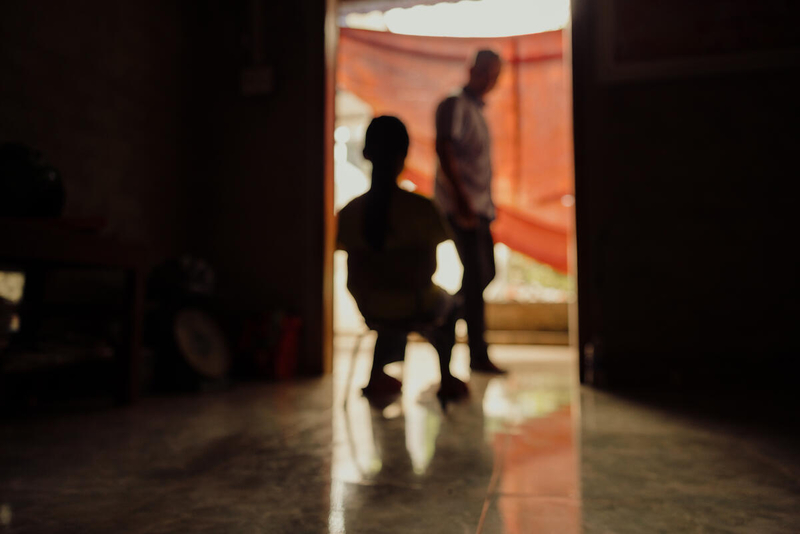 We see the silhouette of a young boy on a chair, in the doorway of a building. A man walks past the doorway. 