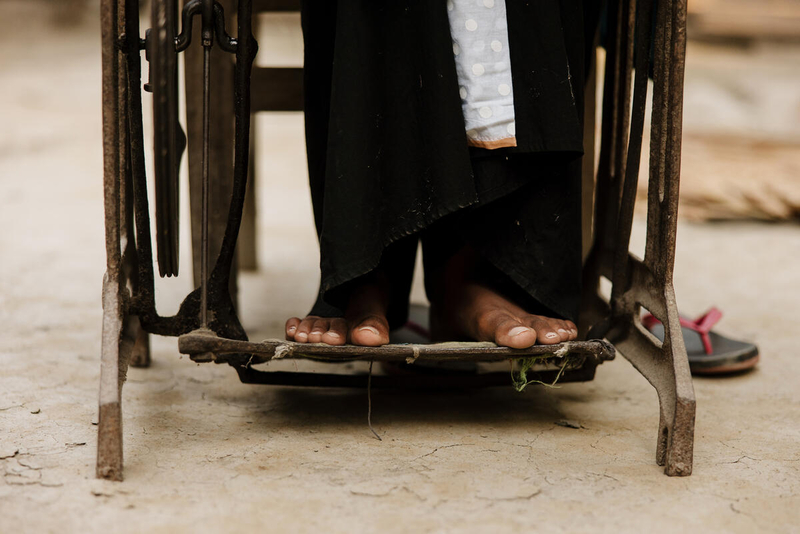 We see the bare feet of a girl on the treadle of a sewing machine. The ground beneath is also bare 