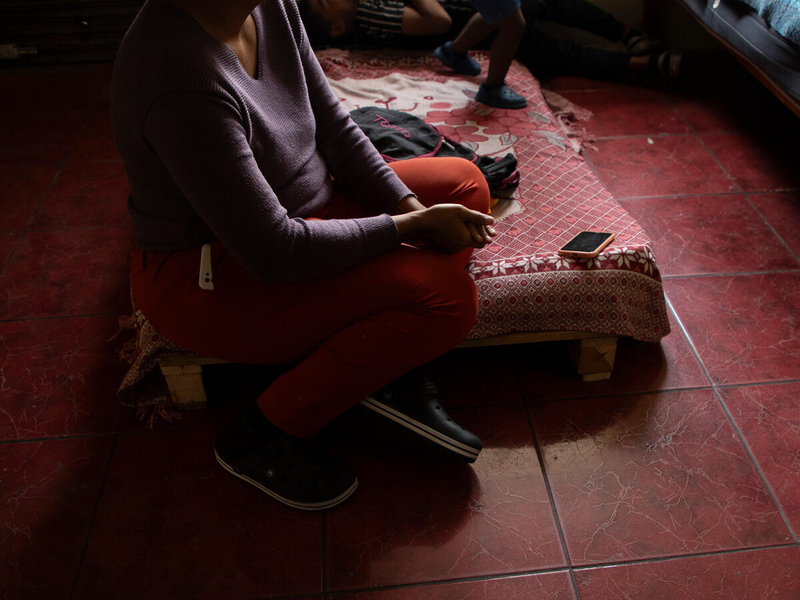 We see an adolescent girl from the neck down, sitting on the edge of a mattress on a tiled floor. 