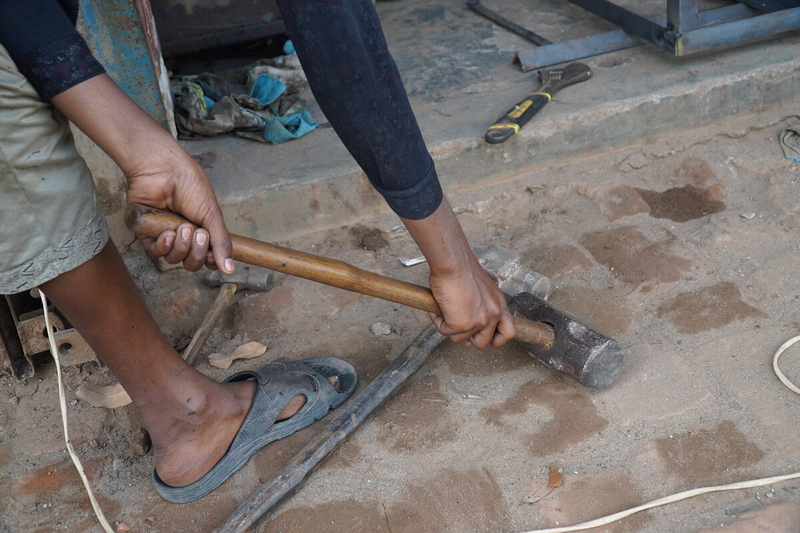 A boy prepares to lift a heavy sledgehammer. We see just his hands, arms, foot and the hammer. 