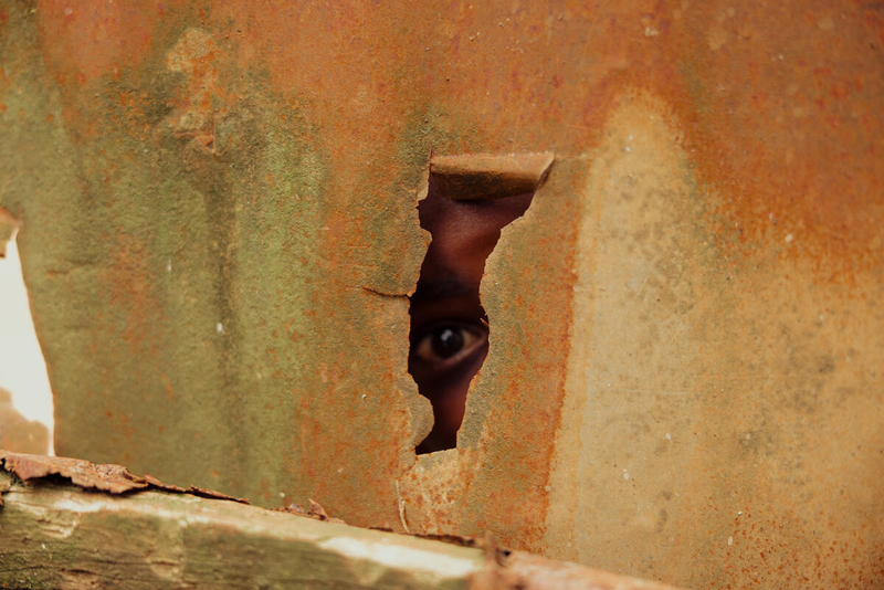 A child peeks through a gap in a wall. We see just one eye and a forehead.