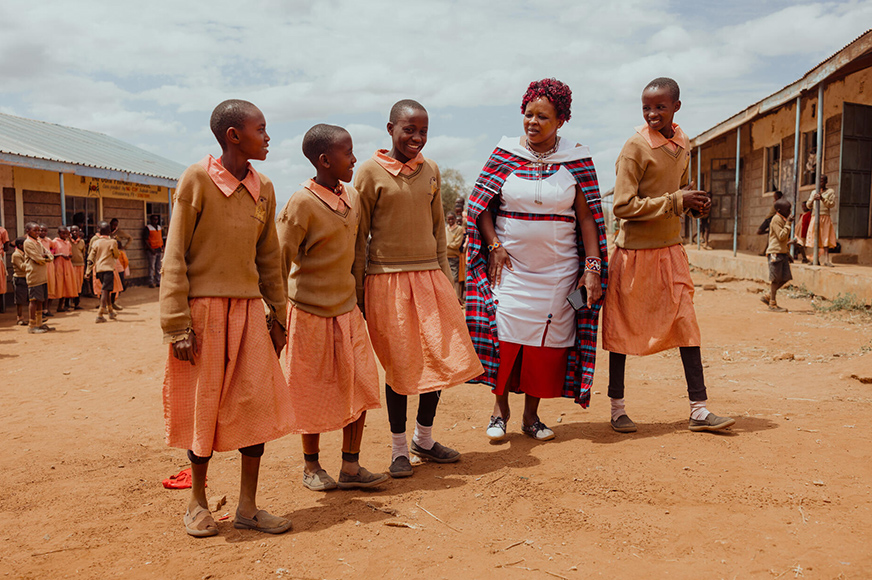 Peninah, wearing a white dress, walks with four students in the schoolyard. She has become a trusted friend and advocate for local girls.