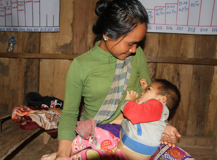 A Laotian woman breastfeeds her child. She is wearing a green shirt and smiling at her child.
