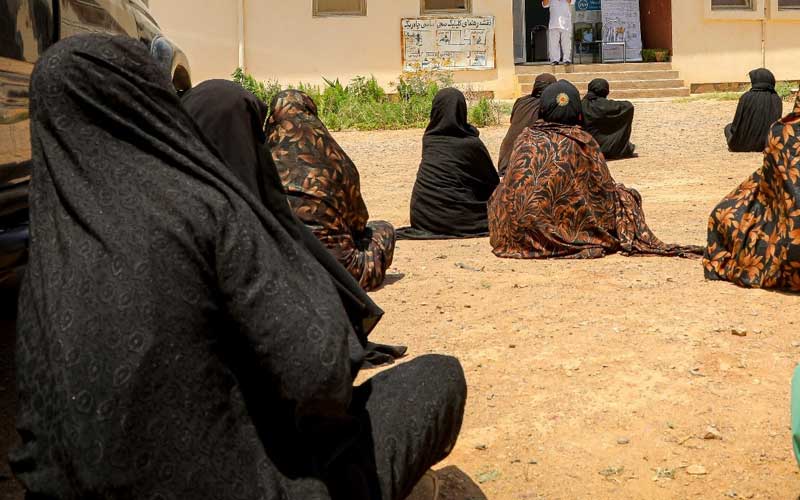 In Afghanistan, a group of women wearing dark head scarves sits on the ground.