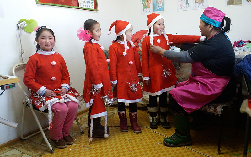 A lady helps four small girls put on red hats that go with their red dresses.