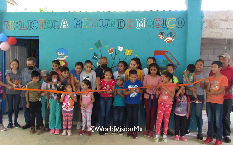 Dozens of children standing in front of a newly established community library in Veracruz, cutting a long ribbon for its inauguration. The library is painted blue with the title “Library - My Magical World”.