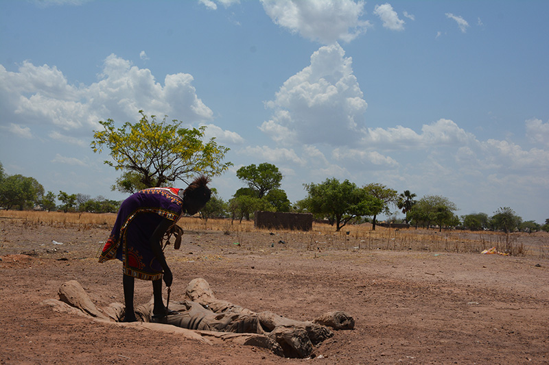 A nineteen-year-old woman bends to scoop water from a ditch in the middle of a parched landscape.