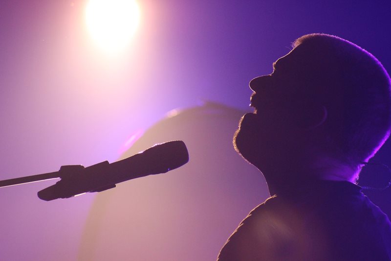 Purple light shines down on a man’s silhouette as he sings in front of a microphone.
