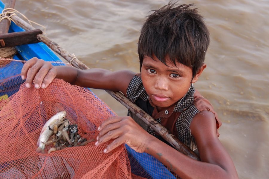 A child labourer in the water holds a net containing fish over the edge of a boat.